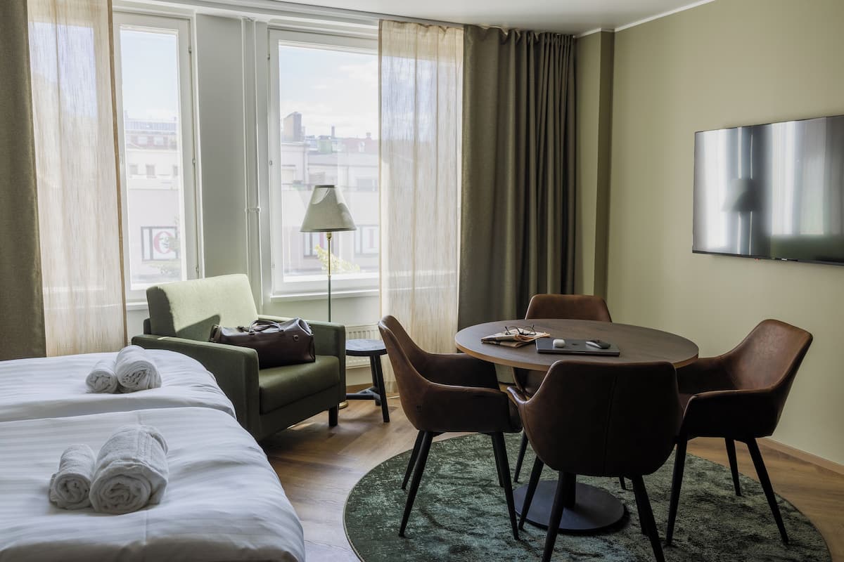 A hotel room with two beds, a dining table with chairs, a sofa, a TV, and a view of the city through large windows.