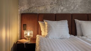 A neatly made bed with white pillows and a striped blanket, a bedside table with a book and a lamp, and a textured wall behind the bed. - Hotel H28 (Tampere)