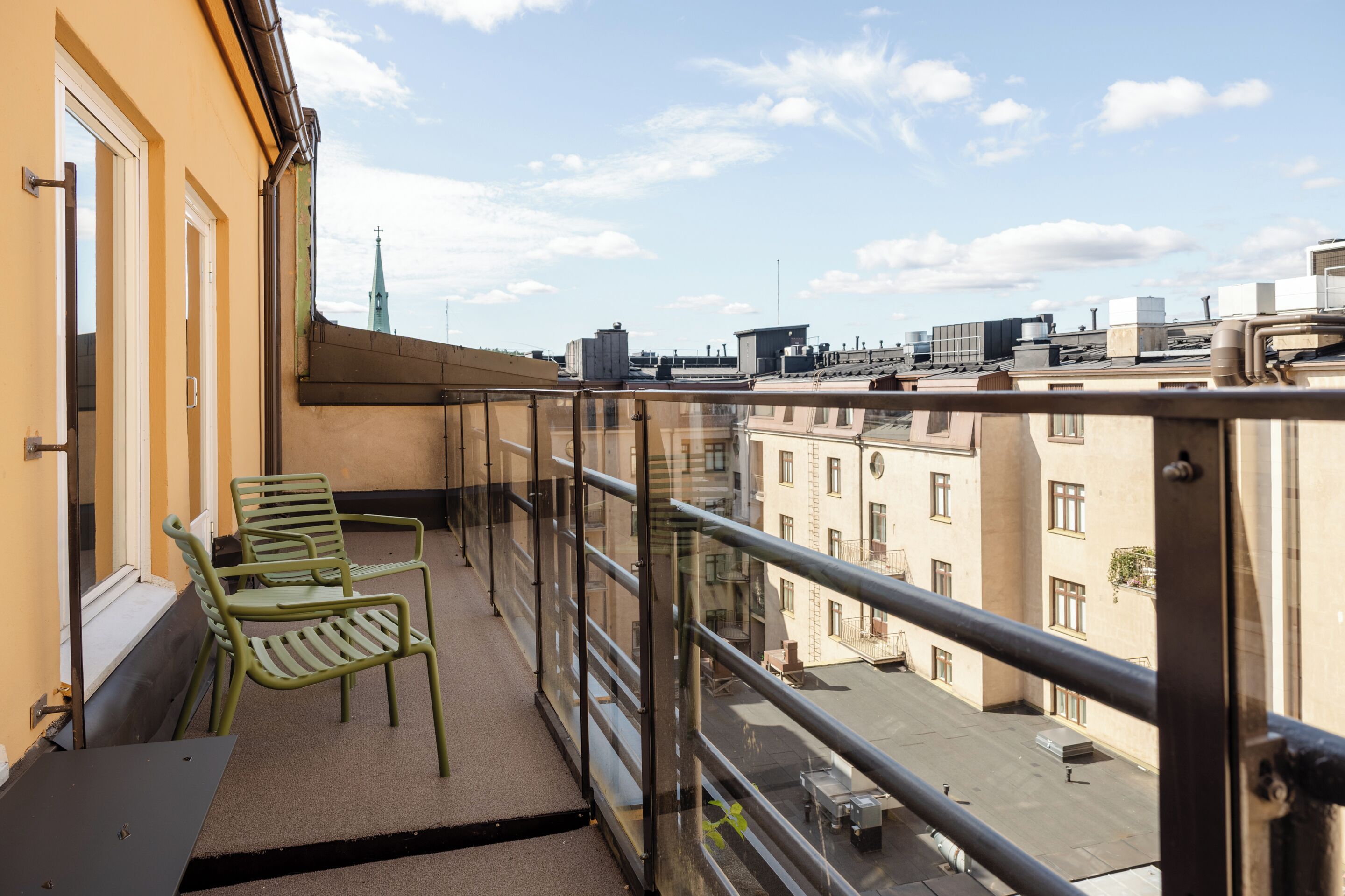 A balcony with two green chairs, a glass railing, and a view of buildings and a church spire.