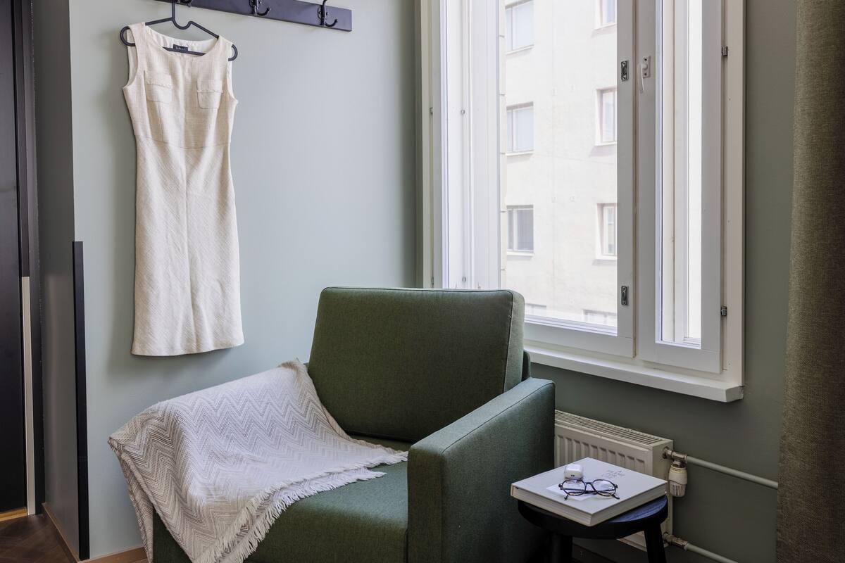 A room with a green armchair, a white dress hanging on a hook, and a window with a view of buildings.