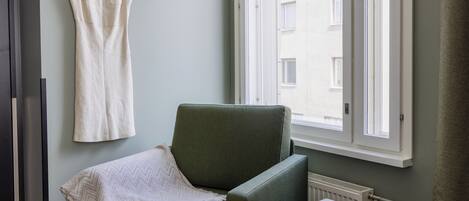 A room with a green armchair, a white dress hanging on a hook, and a window with a view of buildings.