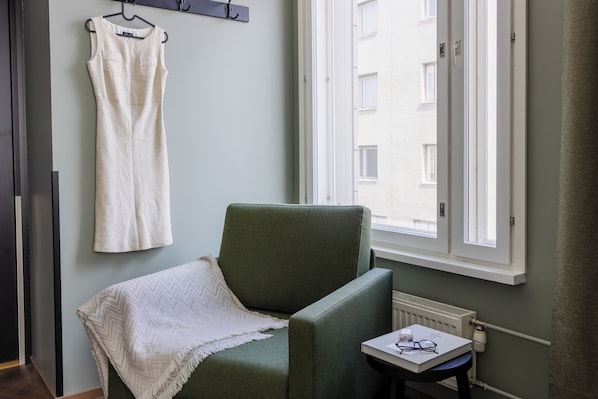 A room with a green armchair, a white dress hanging on a hook, and a window with a view of buildings. - Hotel H28 (Tampere)