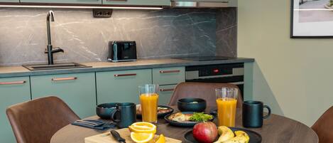 A modern kitchen with a round dining table, a toaster, and breakfast items including orange juice, a banana, and an apple.