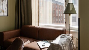 A room with a brown leather sofa, a chair, a lamp, and a view of buildings through a window. - Hotel H28 (Tampere)