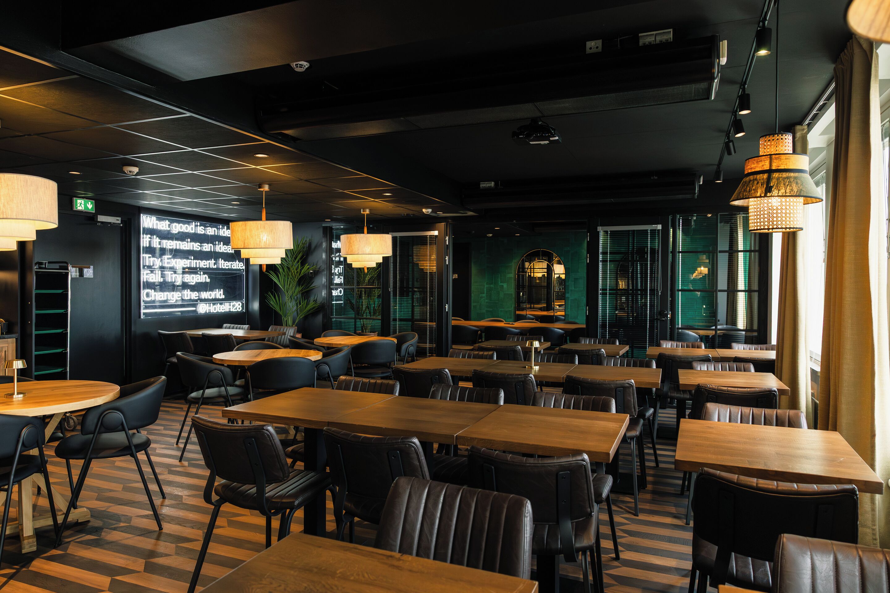 Um restaurante moderno com mesas de madeira e cadeiras pretas, uma tela grande exibindo texto e iluminação pendente.