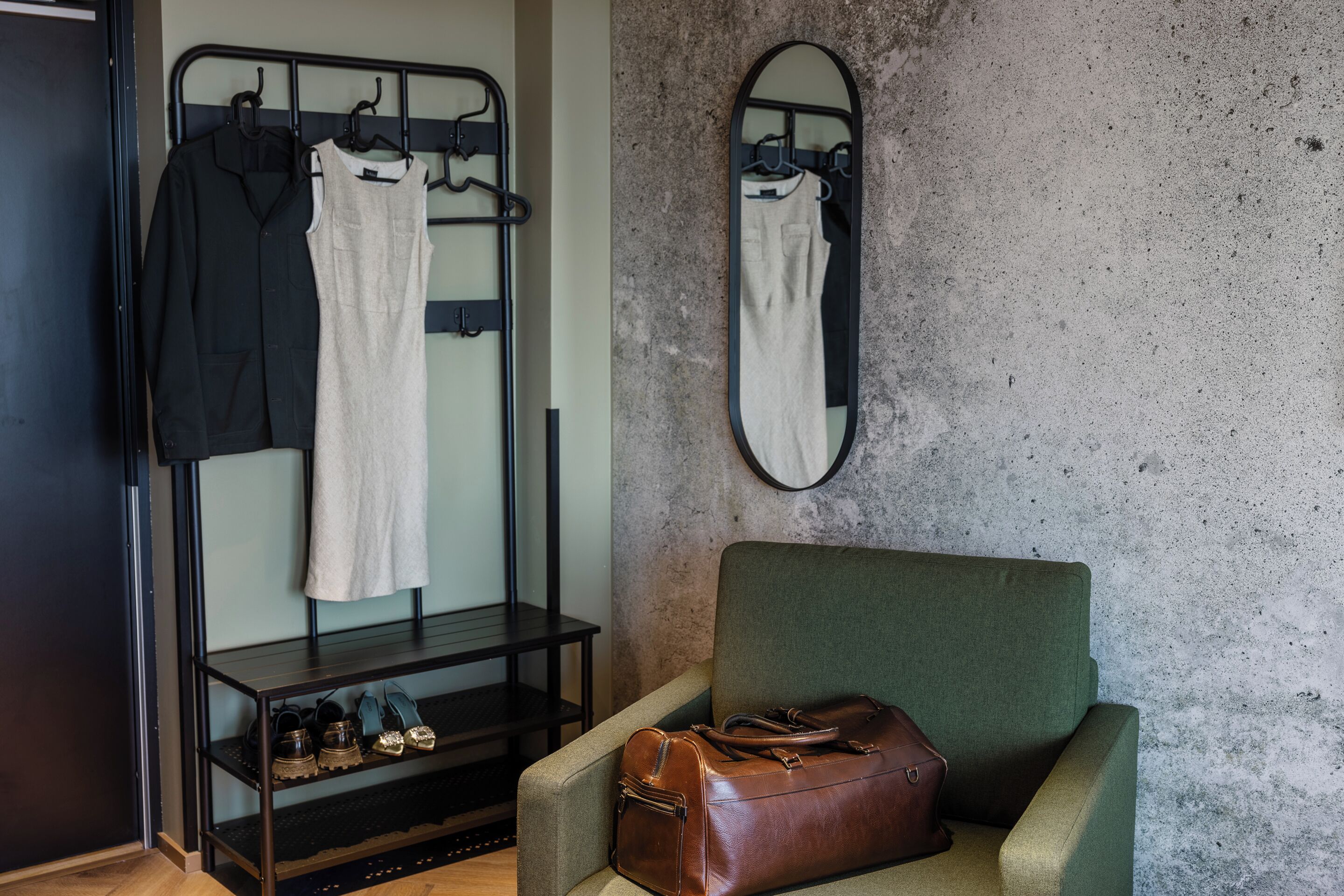 A room with a green armchair, a brown leather bag, a metal rack with clothes, and a mirror on the wall.