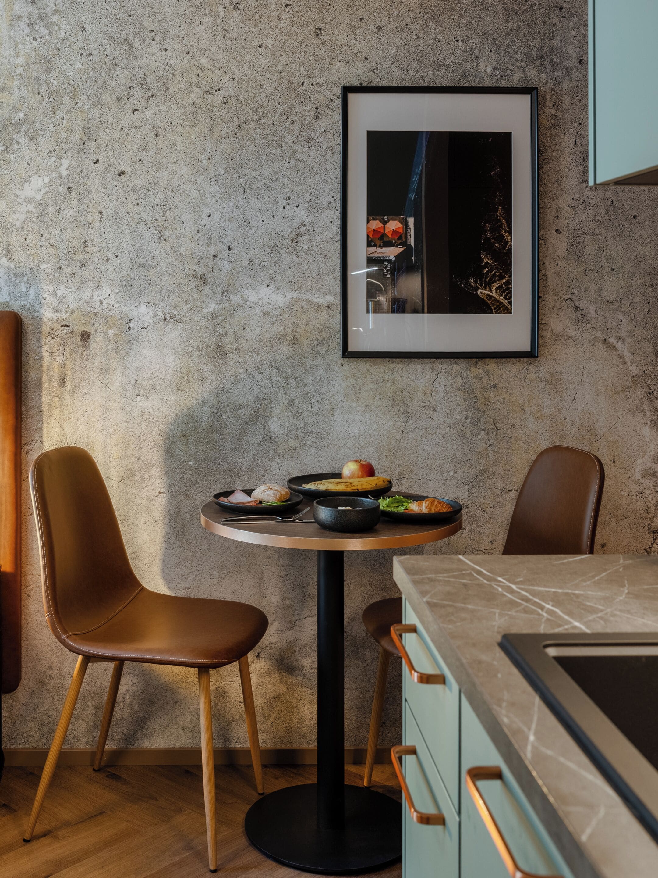 Un comedor moderno con una mesa redonda, dos sillas de cuero, un cuadro en la pared y una encimera de cocina con cajones.