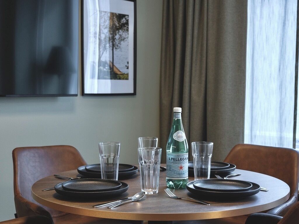 A round dining table with black plates, clear glasses, cutlery, and a bottle of Pellegrino water.