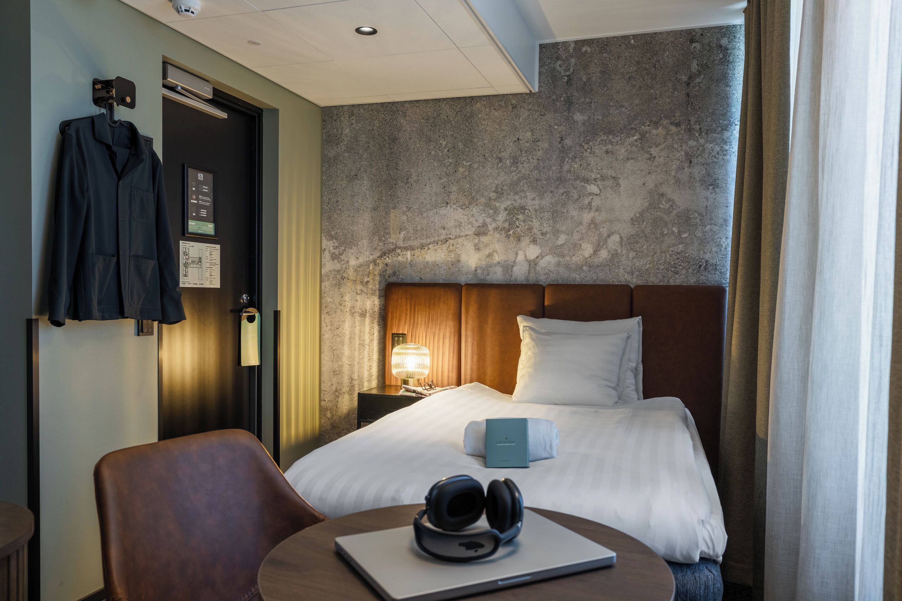 Ein Hotelzimmer mit Bett, Schreibtisch mit Kopfhörern, Stuhl, Kleiderständer und Nachttisch mit Lampe.