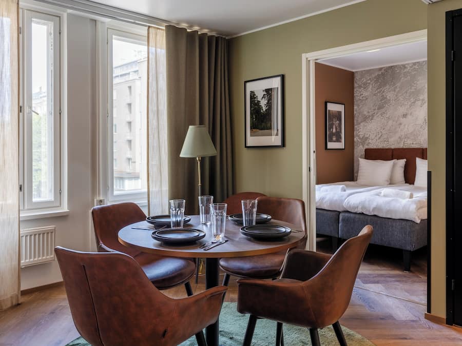 A hotel room with a dining area featuring a round table, brown leather chairs, and a lamp. There is a bed with white bedding and a headboard, and a framed picture on the wall.