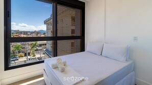 A bedroom with a large window, a bed with white linens, and a view of buildings and a mountain. - Balcony With sea View 230m From the Beach! Prb0405 (Florianópolis)