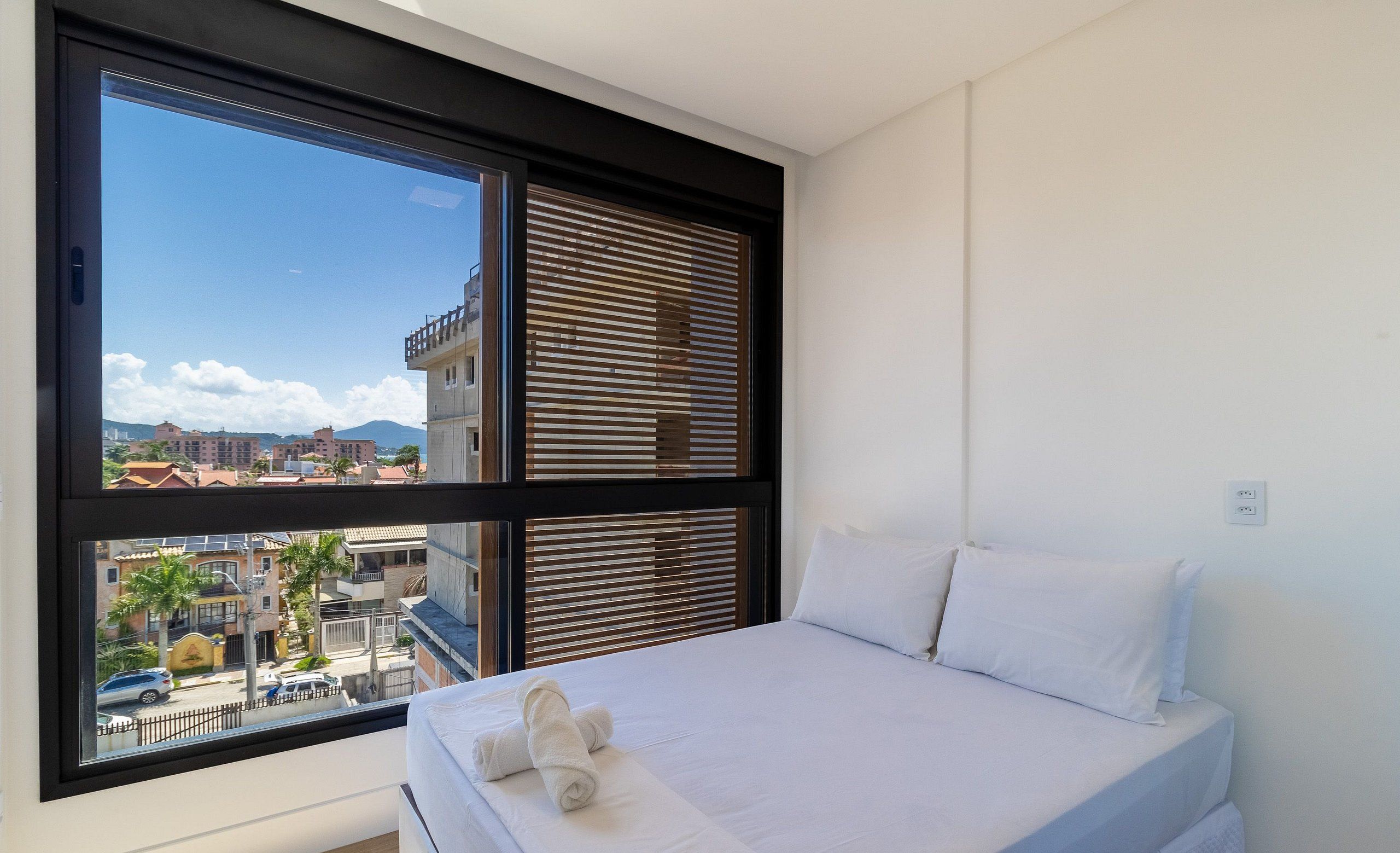 A bedroom with a large window, a bed with white linens, and a view of buildings and a mountain.