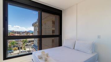 A bedroom with a large window, a bed with white linens, and a view of buildings and a mountain.