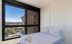 A bedroom with a large window, a bed with white linens, and a view of buildings and a mountain. - Balcony With sea View 230m From the Beach! Prb0405 (Florianópolis)