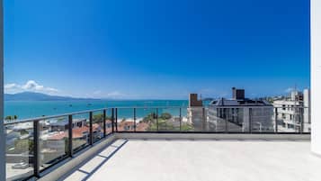 A balcony with a clear view of the sea and boats, a glass railing, and a tiled floor.