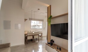 Un salón moderno con un televisor de pantalla plana montado en una pared de madera, una zona de comedor con mesa y sillas, y un ventanal que proporciona luz natural.