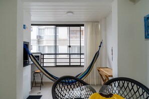 Egy szoba, amelyben van egy függőágy, egy kis faasztal, egy fekete szék, és kilátás nyílik egy fehér korláttal ellátott erkélyre.