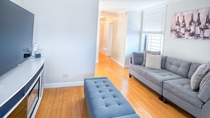 A modern living room with a flat-screen TV, a grey sofa, a blue ottoman, and a painting on the wall. - Columbia University 3Bedroom 1 1/2 Bath Across Cathedral of St. John the Divine (New York)