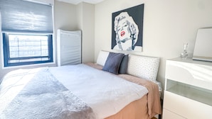 A bedroom with a bed, a bedside table, a window with blinds, and a wall with a pop art painting. - Columbia University 3Bedroom 1 1/2 Bath Across Cathedral of St. John the Divine (New York)