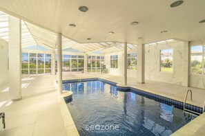 Ein Hallenbad mit klaren Glaswänden, gefliestem Boden und einer Decke mit eingelassenen Leuchten.