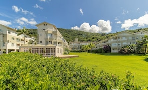 A residential building complex with multiple balconies, a well-maintained lawn, and a mountainous backdrop. - Apartamento na Lagoa da Conceicao Plg0012 (Florianópolis)
