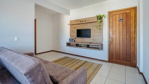 A living room with a sofa, a TV mounted on a wooden wall, a door, and a plant. - Apartment With Balcony in Lagoa Pir0014 (Florianópolis)