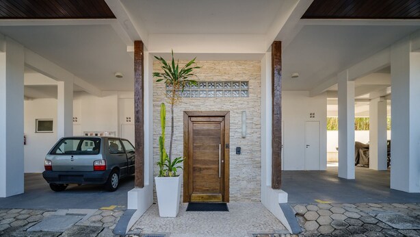 A modern building entrance with a stone wall, a wooden door, and a car parked outside. - Apartment With Balcony in Lagoa Pir0014 (Florianópolis)