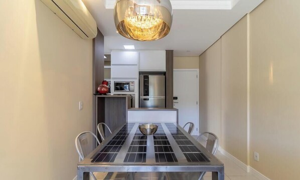 Uma sala de jantar com uma mesa, cadeiras, um candelabro e uma cozinha ao fundo.
