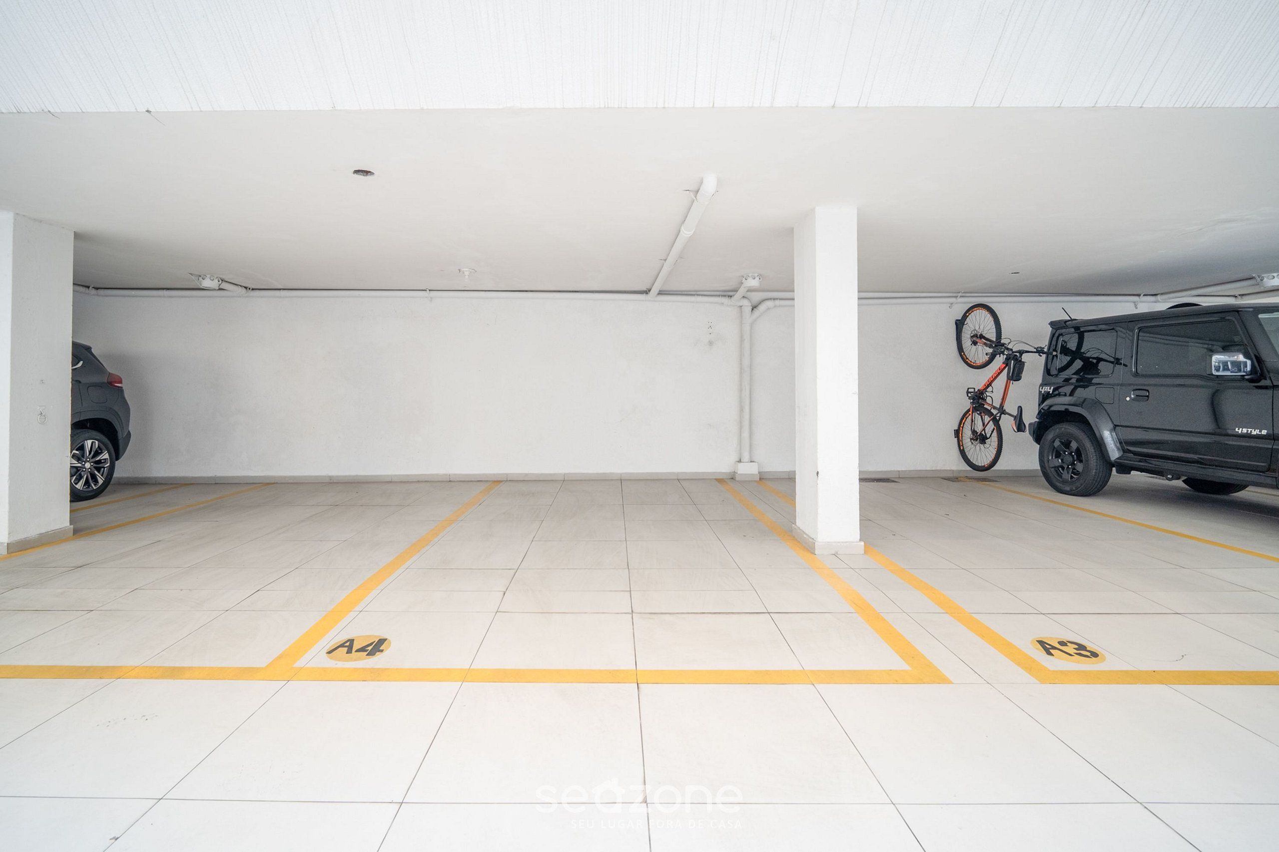 Un aparcamiento con paredes blancas y suelo de baldosas, plazas de aparcamiento marcadas A4 y A3, y una bicicleta colgada en una pared.