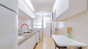 Uma cozinha estreita com armários brancos, uma pia de aço inoxidável e uma pequena mesa de jantar com uma cadeira.