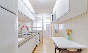 A narrow kitchen with white cabinets, a stainless steel sink, and a small dining table with a chair. - Apt Floripa, 50m to the Beach - Pad0103 (Florianópolis)