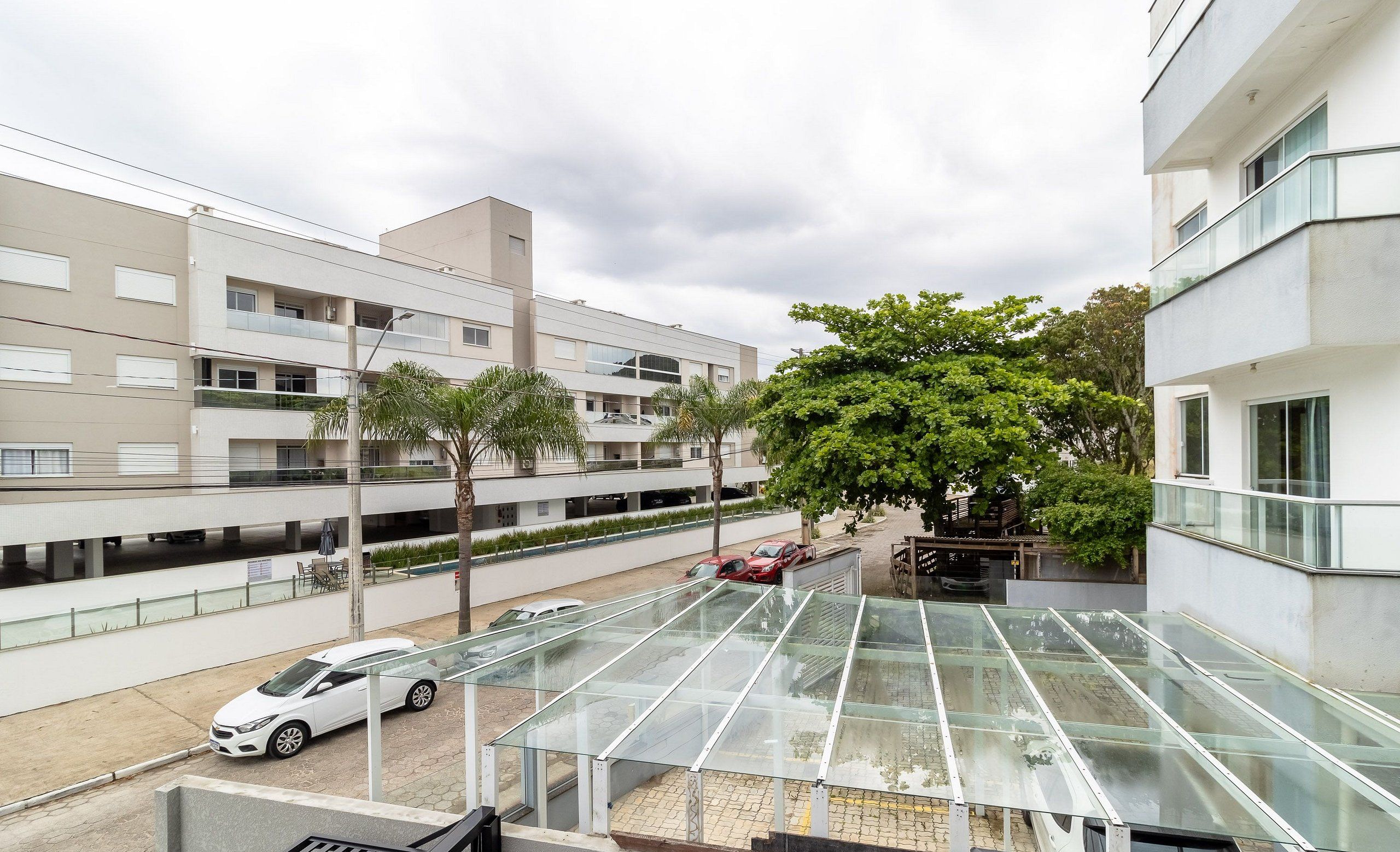 Um edifício residencial moderno com área externa envidraçada, um carro branco estacionado nas proximidades e uma árvore oferecendo sombra.