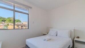 A bedroom with a large window, a bed with white linens, a nightstand, and a view of buildings outside. - Cozy Apartment in Capoeiras, Floripa Opt504 (Florianópolis)