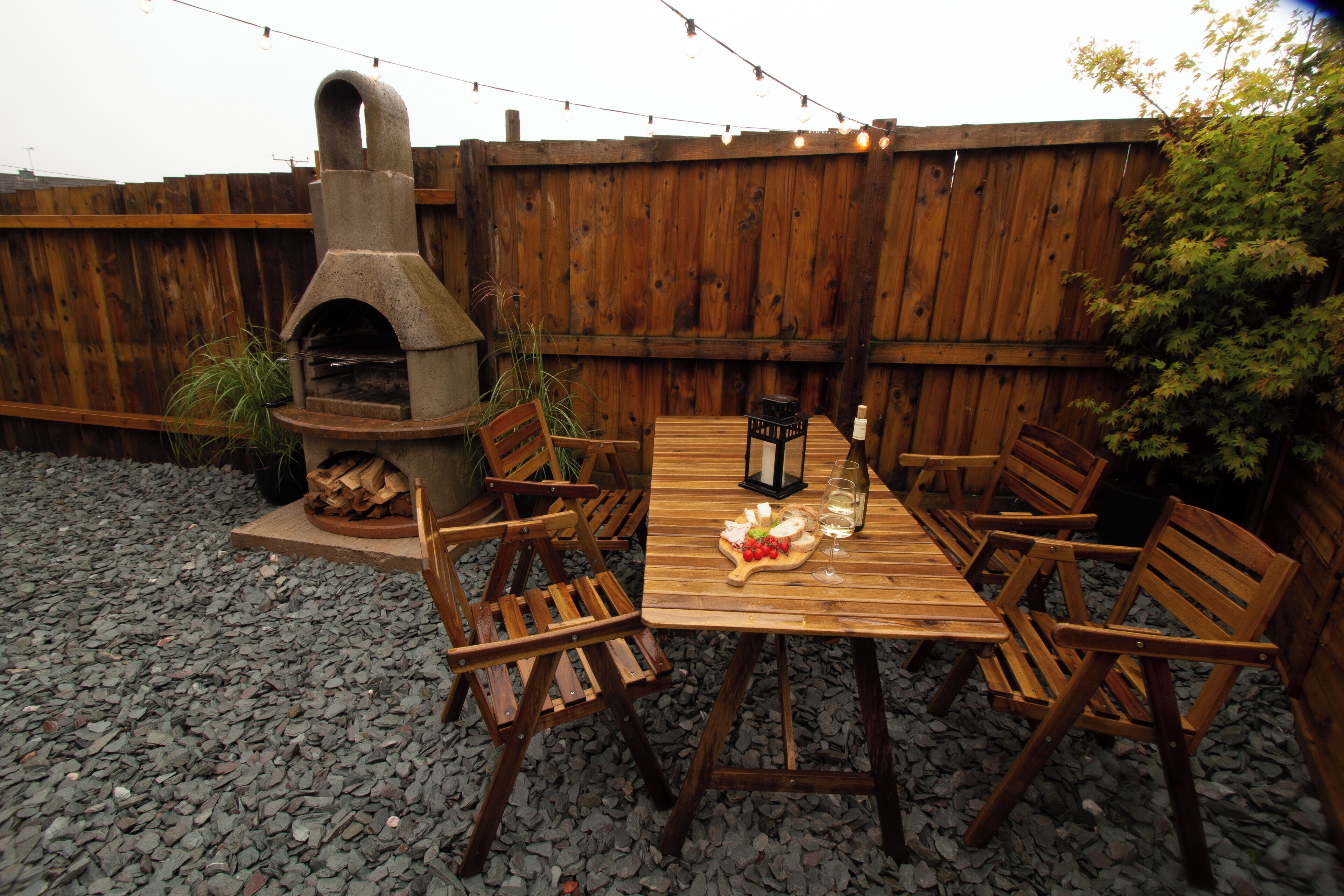 An outdoor patio with a wooden table and chairs, a stone oven, and a string of lights.