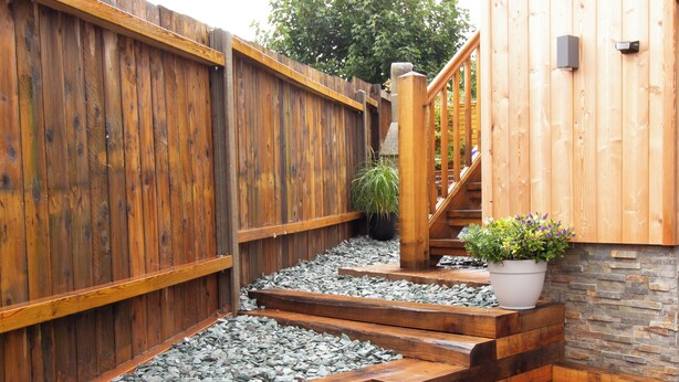 A wooden deck with steps leading to a stone wall, a wooden fence, and a staircase with a railing.