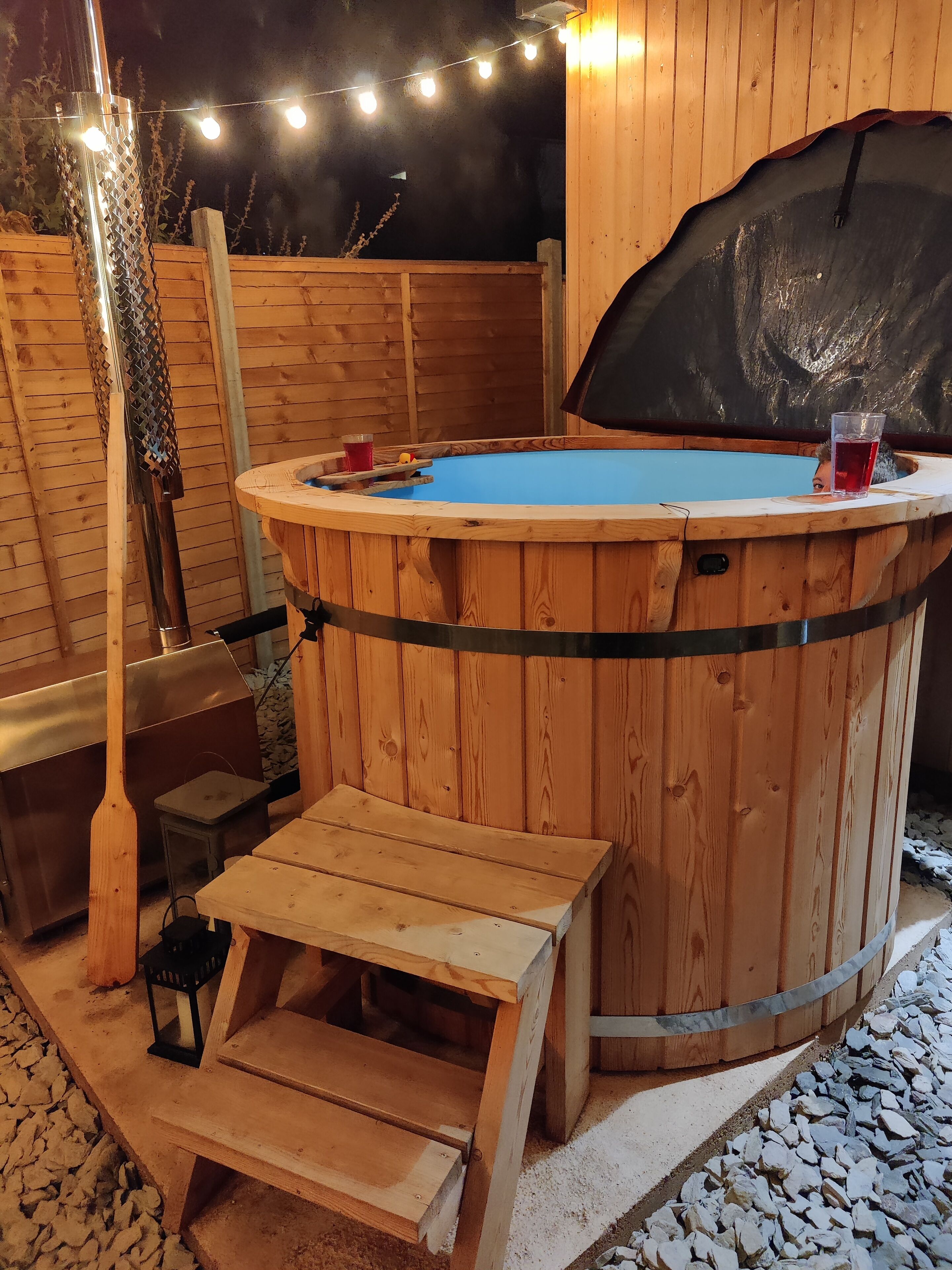 A wooden hot tub with a wooden deck and steps leading into it, surrounded by a wooden fence and string lights.