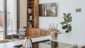 Un salon moderne avec une bibliothèque en bois, un téléviseur à écran plat, une grande plante et un coin repas comprenant une table et des chaises.