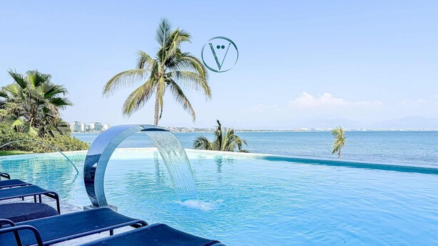 Una piscina con fuente, sillas de descanso, palmeras y vista al mar.
