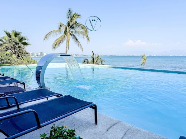 A pool with a water feature, lounge chairs, palm trees, and a view of the sea.