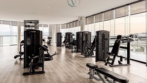 Un gimnasio con diversas máquinas de ejercicio, amplias ventanas y vistas a la ciudad.