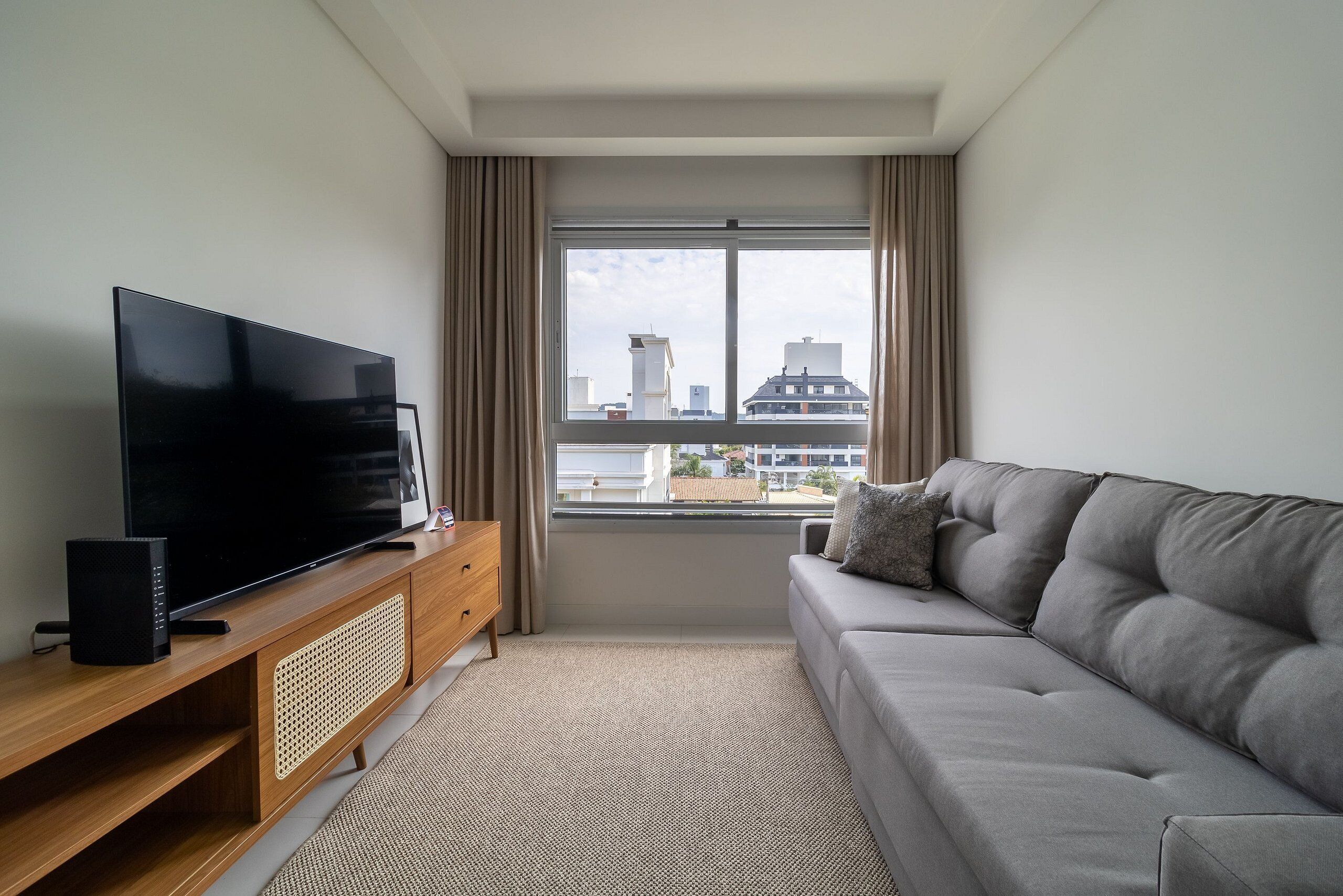 A modern living room with a flat-screen TV on a wooden stand, a grey sofa, and a large window with curtains.