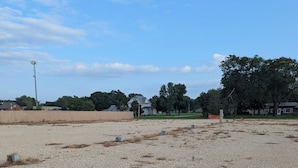 A gravel parking area with sparse vegetation, a utility pole, and residential houses in the background. - Next Door to the Fair RV Park (Spencer)