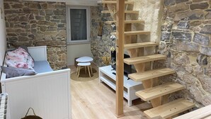 A cozy room with a stone wall, a wooden staircase, a white sofa, and a small round table. - The House of Evahlia (Creissels)