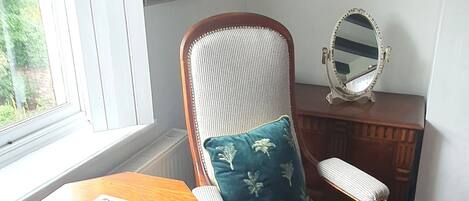 A room with a wooden chair with a cushion, a small wooden table, a mirror, and a framed picture on the wall.