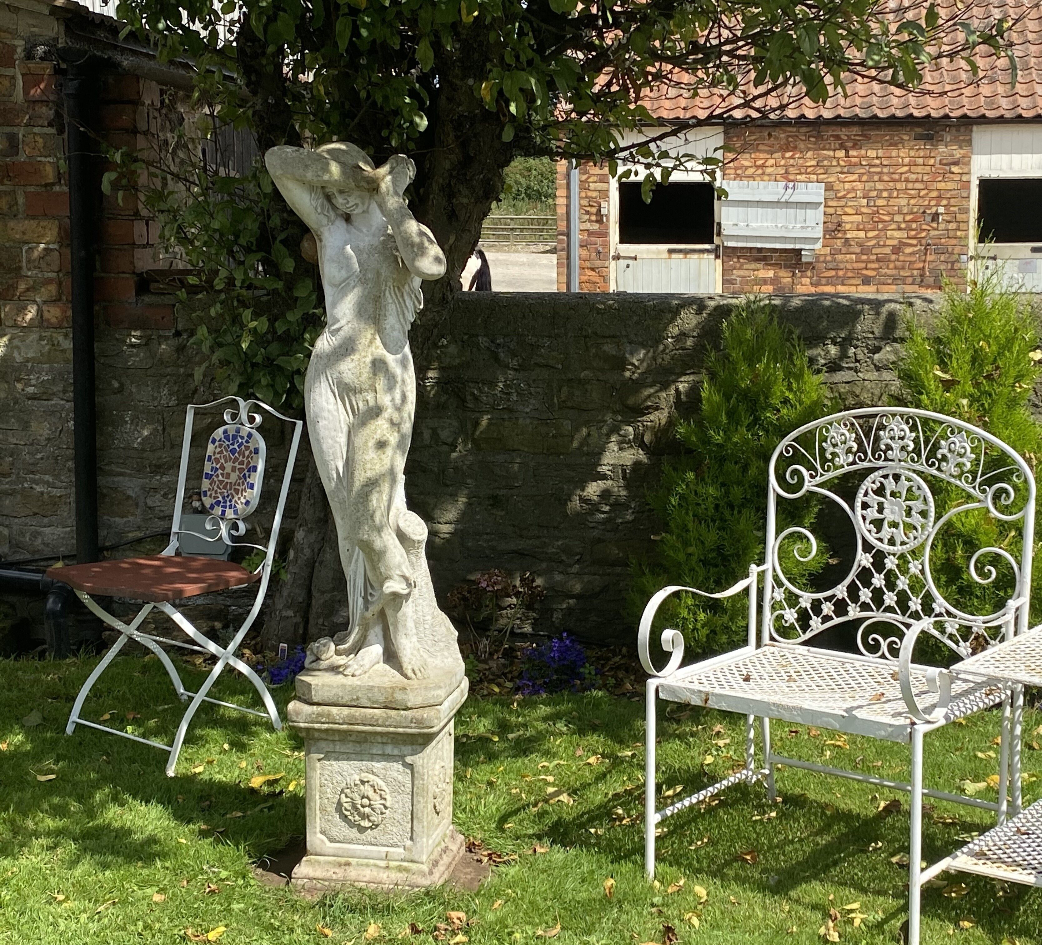 A statue of a woman on a pedestal in a garden with two metal chairs and a stone wall in the background.
