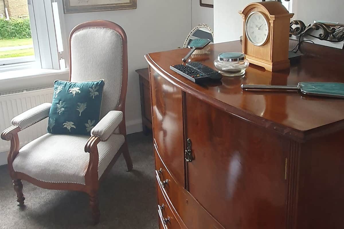 Een kamer met een houten dressoir, een stoel met een blauwe kussentje, een ingelijste kaart en een klok op het dressoir.