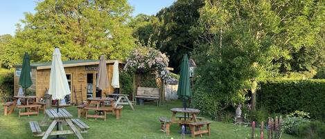 A garden with wooden picnic tables and green umbrellas, surrounded by lush greenery and a wooden building labeled "ANTIQUE".