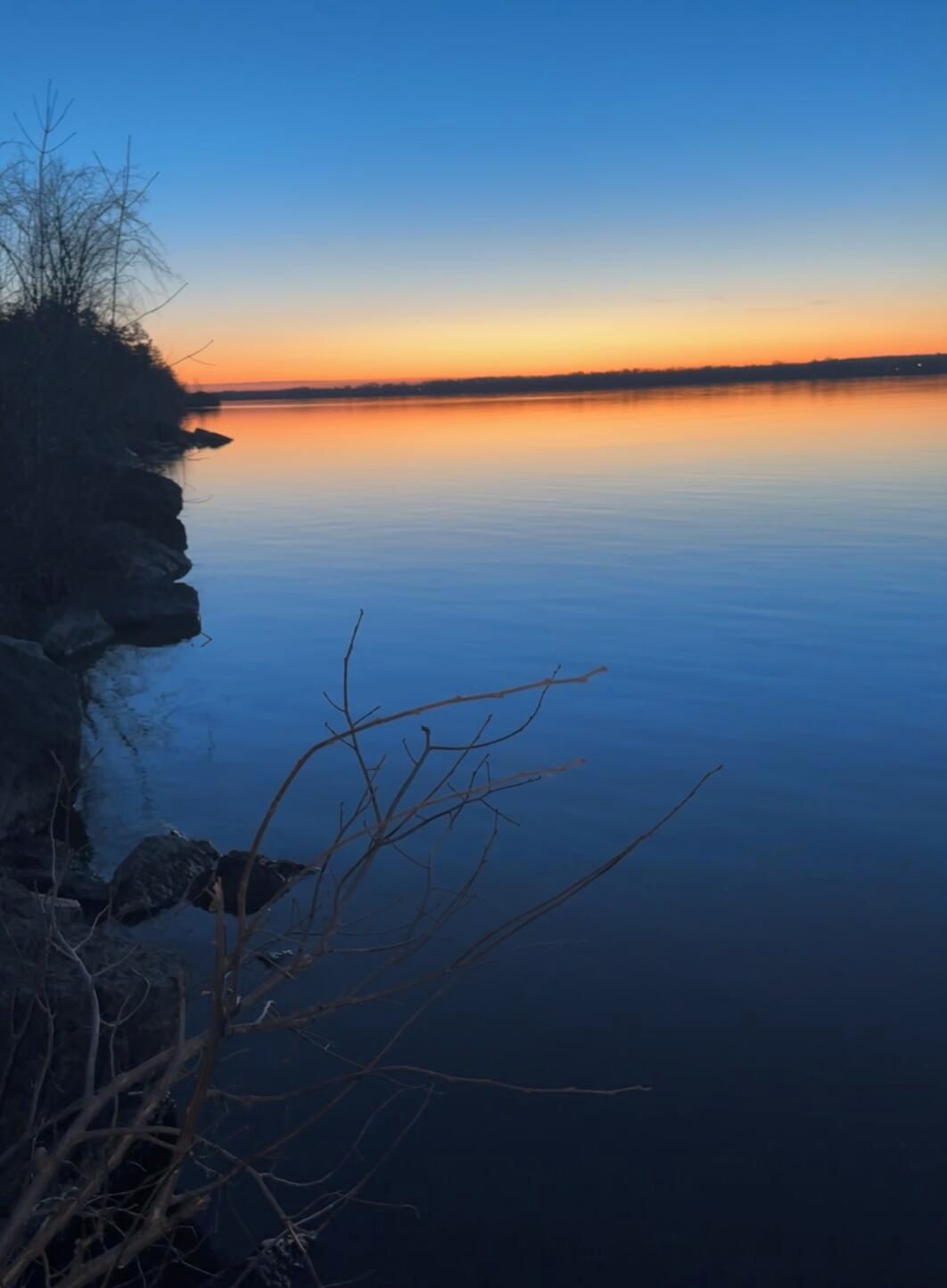 Un lago tranquilo al atardecer con una orilla rocosa y ramas desnudas en primer plano.