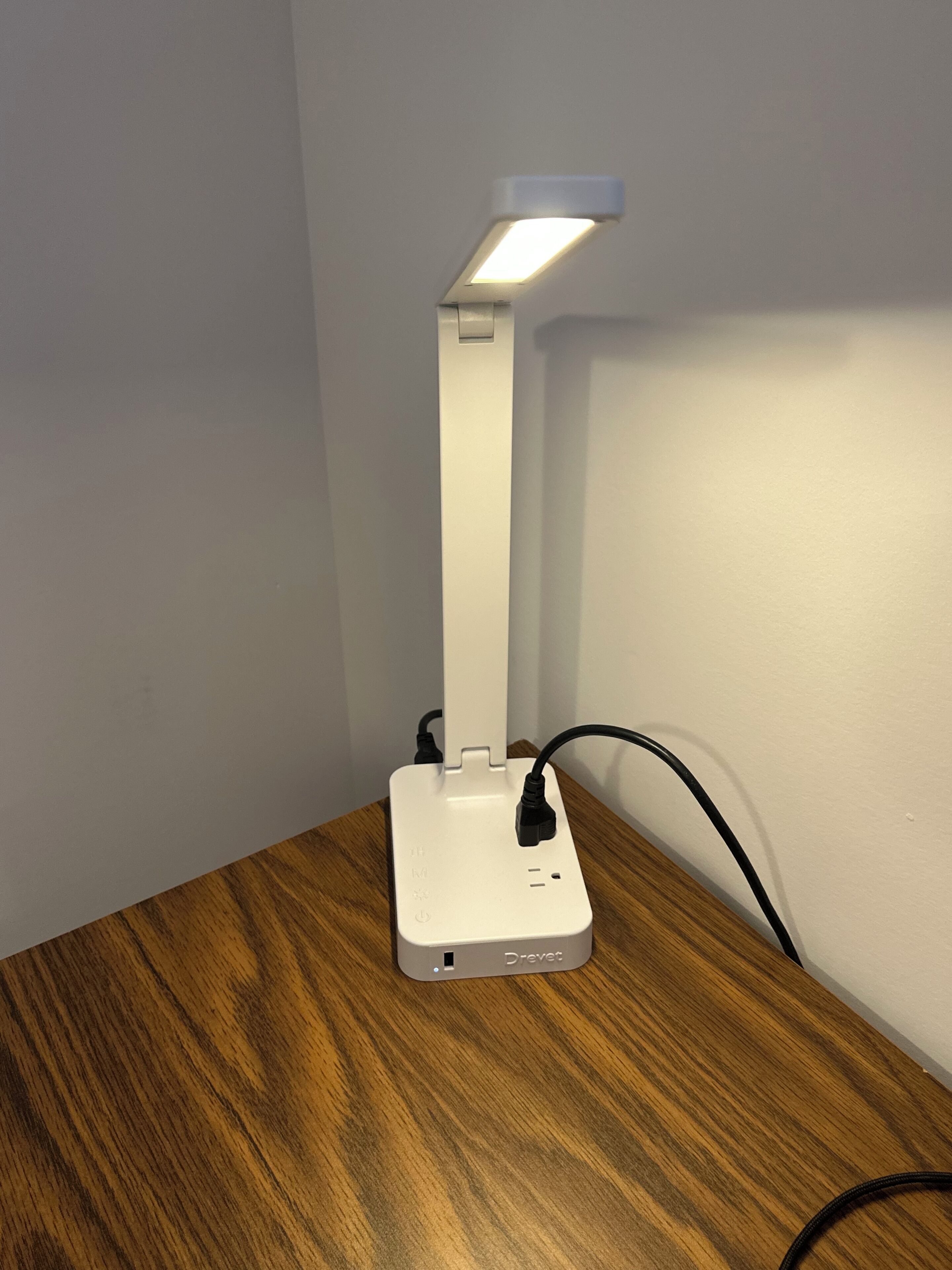 A white desk lamp with a USB port and a power cord on a wooden desk.