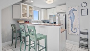 Una cocina con armarios blancos, una isla central con taburetes de bar, un refrigerador y una obra de arte con forma de caballito de mar en la pared.